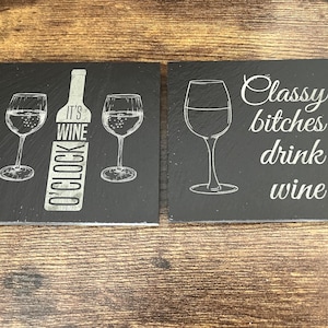 May include: Two black slate coasters with white etched designs. One coaster features two wine glasses and a wine bottle with the text "IT'S WINE O'CLOCK". The other coaster says "Classy bitches drink wine".