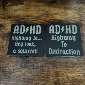 May include: Two black coasters with white text. The first coaster says "AD HD Highway To... Hey look, a squirrel!" The second coaster says "AD HD Highway To Distraction".
