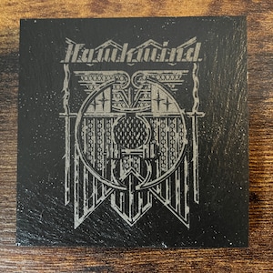 May include: Black slate coaster with a silver design featuring the word "Remarkable" and a stylised graphic of a shield with wings.