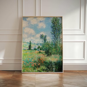 May include: A framed landscape painting featuring a vibrant field of red poppies and green grass under a blue sky with fluffy white clouds. Tall green trees and a distant village are also visible. The artwork is in a light wooden frame.
