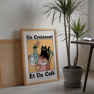 May include: A framed illustration of a black cat sitting at a table with a croissant, a cup of coffee, and a teapot. The text on the illustration reads "Un Croissant Et Un Caf&eacute;".