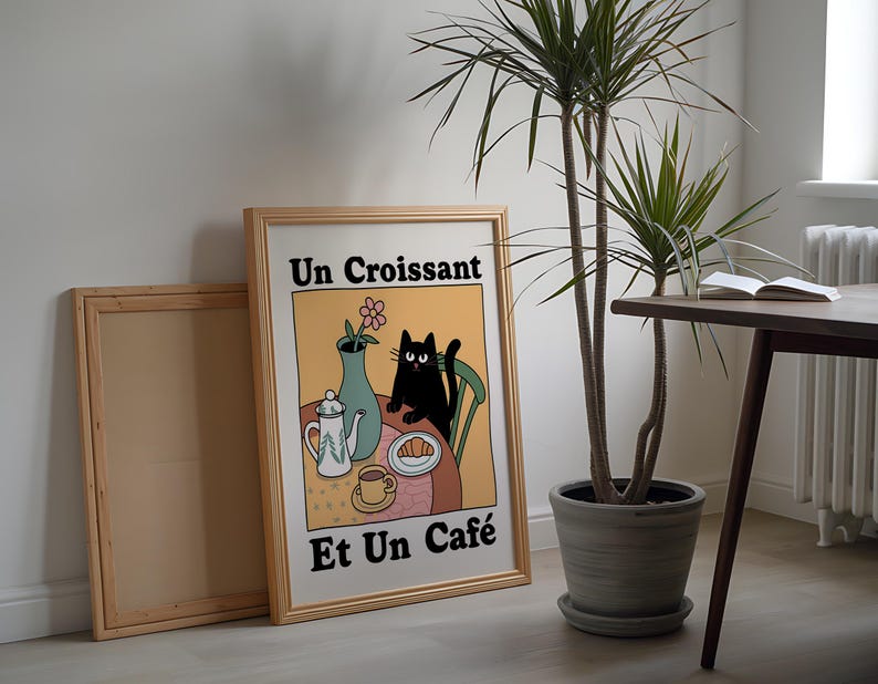 May include: A framed illustration of a black cat sitting at a table with a croissant, a cup of coffee, and a teapot. The text on the illustration reads "Un Croissant Et Un Caf&eacute;".