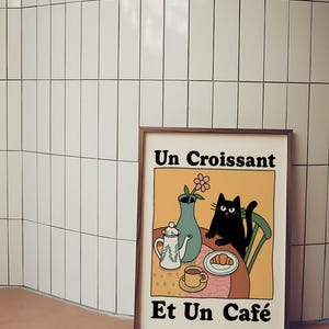 May include: A framed illustration of a black cat sitting at a table with a croissant, a cup of coffee, a teapot, and a vase of flowers. The text on the illustration reads "Un Croissant Et Un Caf&eacute;".