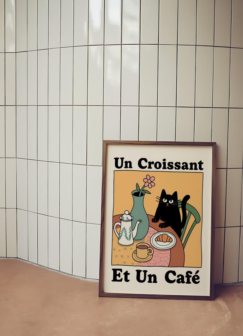 May include: A framed illustration of a black cat sitting at a table with a croissant, a cup of coffee, a teapot, and a vase of flowers. The text on the illustration reads "Un Croissant Et Un Caf&eacute;".