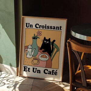 May include: A framed illustration of a black cat sitting on a chair at a table with a teapot, cup, saucer, croissant, and a vase with a pink flower. The text on the illustration reads "Un Croissant Et Un Caf&eacute;".