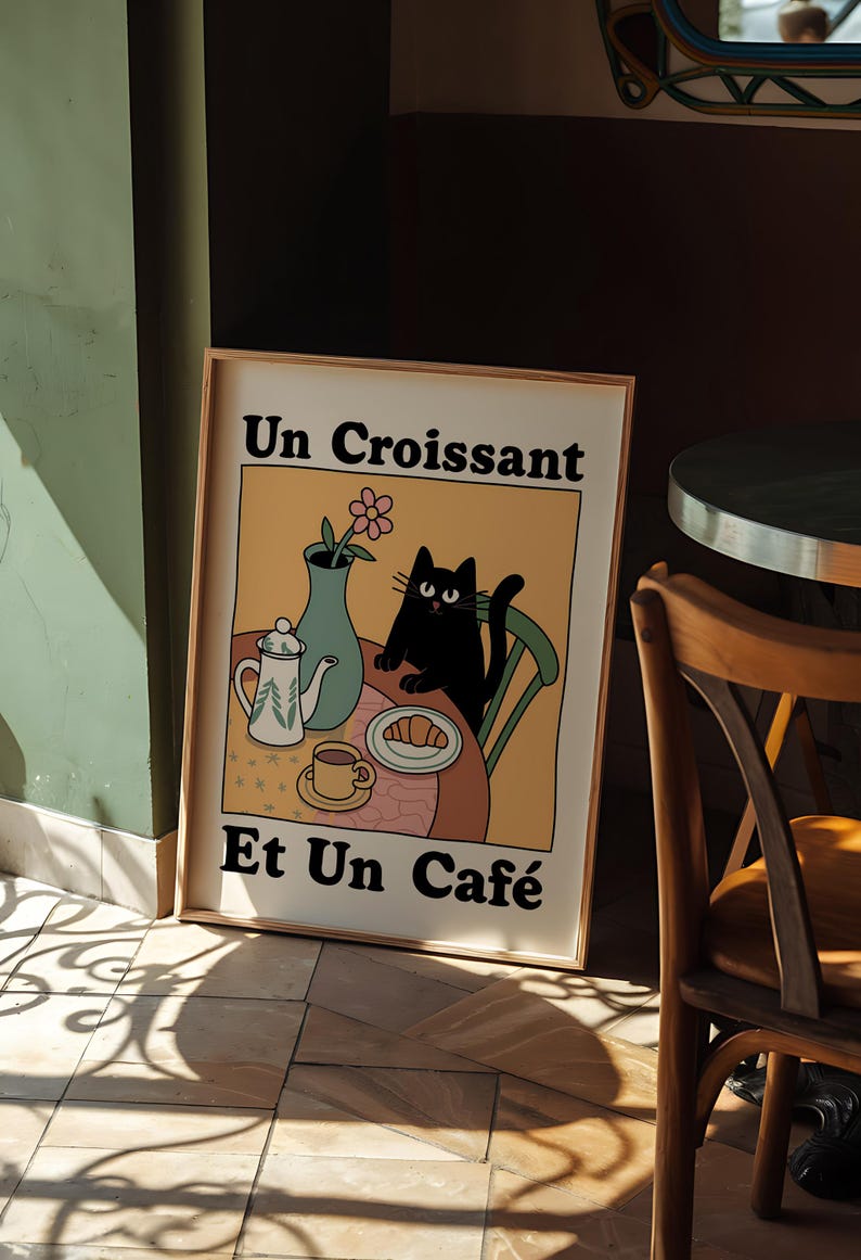 May include: A framed illustration of a black cat sitting on a chair at a table with a teapot, cup, saucer, croissant, and a vase with a pink flower. The text on the illustration reads "Un Croissant Et Un Caf&eacute;".