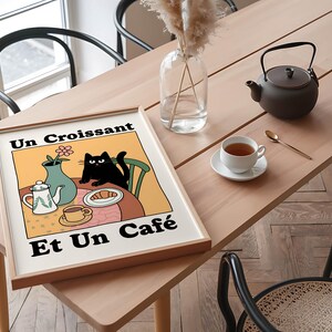 May include: A framed print with a yellow background and a black cat sitting at a table with a croissant, a teapot, and a cup of coffee. The text on the print reads "Un Croissant Et Un Cafe".