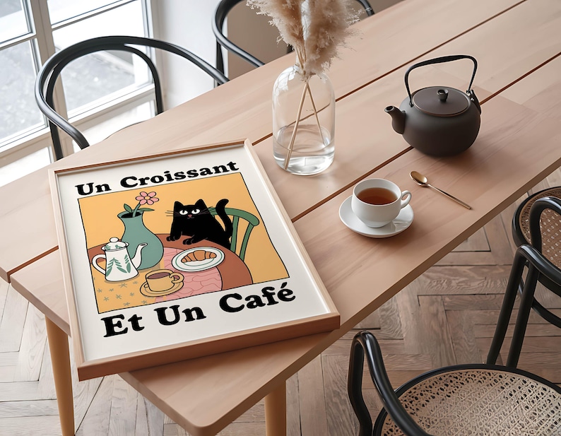 May include: A framed print with a yellow background and a black cat sitting at a table with a croissant, a teapot, and a cup of coffee. The text on the print reads "Un Croissant Et Un Cafe".