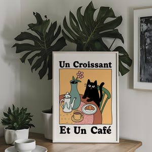 May include: A white framed print with a black cat sitting at a table with a croissant, a cup of coffee, and a teapot. The text on the print reads "Un Croissant Et Un Caf&eacute;".