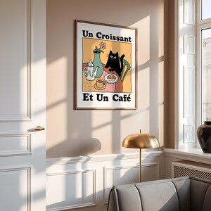 May include: A black cat sits at a table with a cup of coffee, a croissant, and a teapot. The text on the poster reads "Un Croissant Et Un Caf&eacute;".
