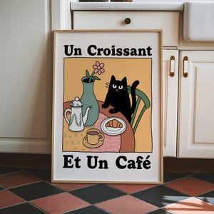 May include: A framed illustration of a black cat sitting at a table with a croissant, a cup of coffee, and a teapot. The text on the illustration reads "Un Croissant Et Un Caf&eacute;".