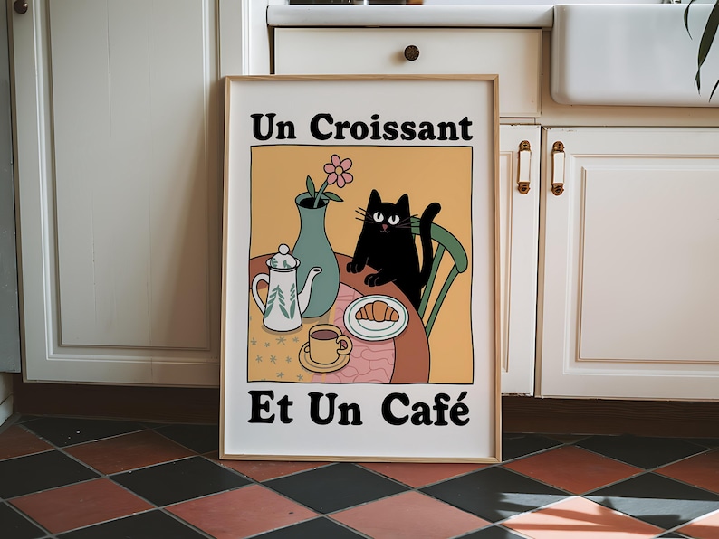 May include: A framed illustration of a black cat sitting at a table with a croissant, a cup of coffee, and a teapot. The text on the illustration reads "Un Croissant Et Un Caf&eacute;".