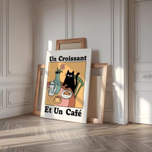 May include: A white poster with a black cat sitting at a table with a croissant, a cup of coffee, and a teapot. The poster has the text "Un Croissant Et Un Caf&eacute;" in black.