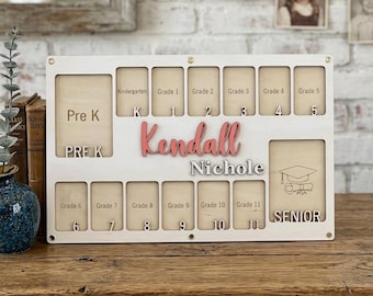 May include: A personalized wooden school years photo display board. The white frame has slots for photos from Pre-K to Senior year. The name "Kendall Nichole" is displayed in pink and silver lettering. The board is on a wooden surface.