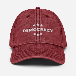Puede incluir: Una gorra de béisbol de algodón lavado vintage de color rojo con bordado blanco que dice "DEMOCRACY" con cinco estrellas arriba y abajo de la palabra.