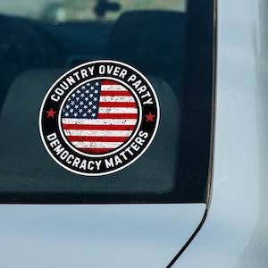 May include: A black and white sticker with a distressed American flag design. The sticker reads "Country Over Party Democracy Matters".