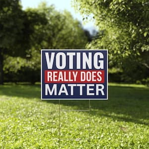 May include: A rectangular yard sign with the text "VOTING REALLY DOES MATTER" in white letters on a navy blue background, with a red stripe in the middle. The sign is held up by a metal wire frame and placed in a grassy yard.