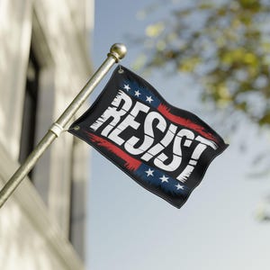Puede incluir: Una bandera negra con la palabra "RESIST" en blanco, texto estilizado. La bandera tiene rayas rojas y azules y estrellas blancas, montada en un mástil dorado. La bandera ondea al viento contra un cielo azul.