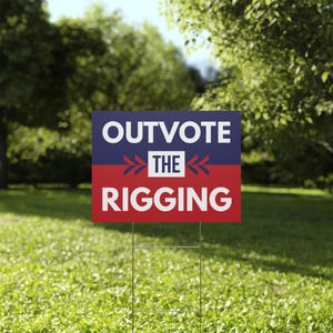 May include: A rectangular yard sign with the text "OUTVOTE THE RIGGING" in white letters. The top half of the sign is blue, and the bottom half is red. The sign is placed in a grassy yard with trees in the background.