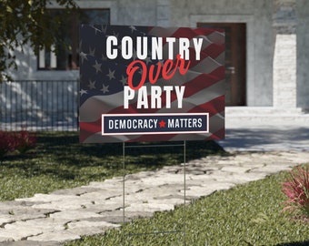 May include: A rectangular yard sign featuring the text "COUNTRY OVER PARTY" in white and red, set against an American flag design. The sign also includes the text "DEMOCRACY MATTERS" in a white box with a red star.
