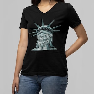 May include: Black V-neck t-shirt featuring a graphic of the Statue of Liberty with her face obscured. The graphic is in shades of gray and blue. The shirt is worn by a person in blue jeans.