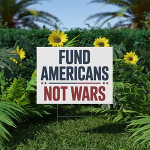 May include: A white yard sign with the text "FUND AMERICANS NOT WARS" in navy blue and red. The sign is supported by a metal frame and set in a grassy area with sunflowers and green foliage in the background.