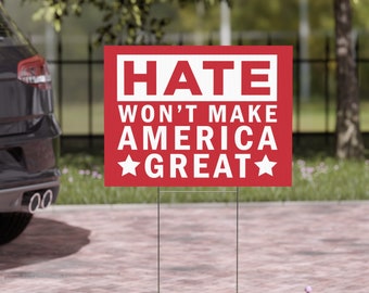 May include: A red yard sign with white text that reads "HATE WON'T MAKE AMERICA GREAT" with white stars. The sign is displayed outdoors, set in grass and a brick paved area, with a car in the background.