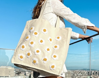May include: A white canvas tote bag with embroidered yellow and white daisies. The bag has a long strap and is being held by a person standing on a balcony overlooking a city.