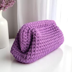 May include: A lavender-colored, handmade clutch bag. The bag is made of thick, woven material, with a rounded, folded top. The bag sits on a white surface, with a white vase of purple flowers in the background.
