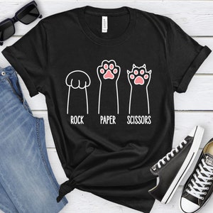 Puede incluir: Camiseta negra con un gr&aacute;fico blanco de una pata de gato, una pata de gato con garras y una pata de gato con un pu&ntilde;o cerrado. El texto "Piedra, Papel, Tijeras" est&aacute; impreso debajo de las patas.