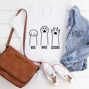 Puede incluir: Camiseta blanca con un gr&aacute;fico de patas de gato jugando a piedra, papel o tijeras. El texto dice "Piedra, papel o tijeras".