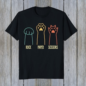 Puede incluir: Camiseta negra con un gr&aacute;fico de tres patas de gato que representan piedra, papel, tijeras. Las patas son de diferentes colores y tienen un dise&ntilde;o retro.