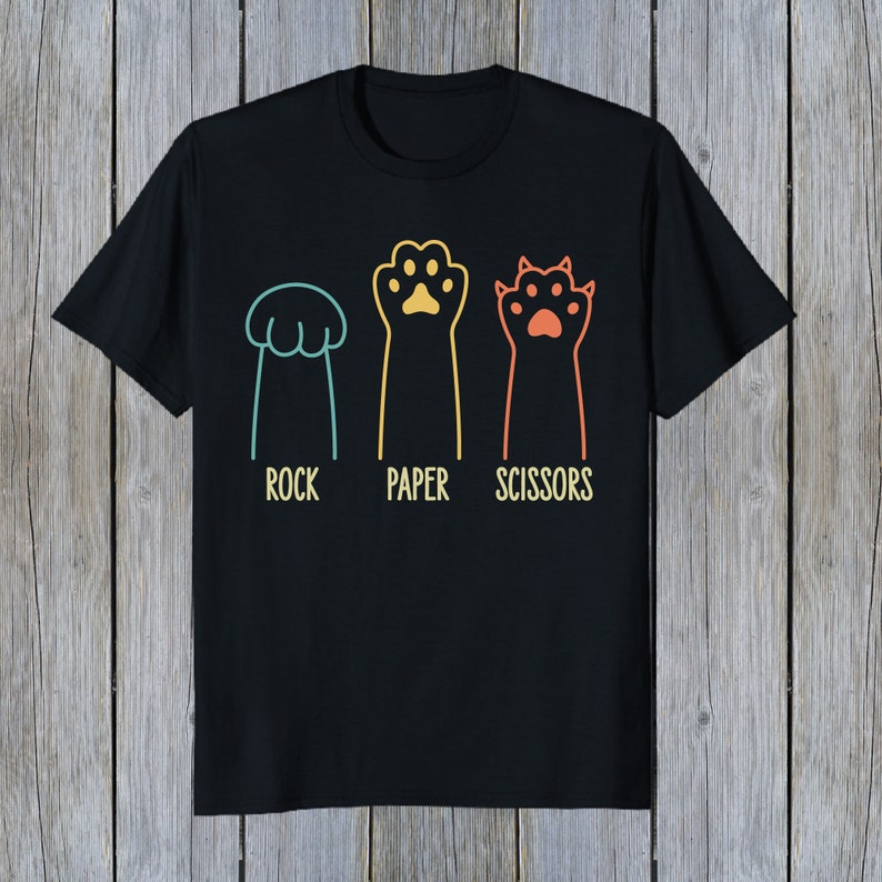 Puede incluir: Camiseta negra con un gr&aacute;fico de tres patas de gato que representan piedra, papel, tijeras. Las patas son de diferentes colores y tienen un dise&ntilde;o retro.