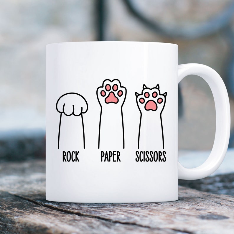 Puede incluir: Taza de cer&aacute;mica blanca con una ilustraci&oacute;n en negro y rosa de una huella de pata, una huella de pata de gato y una huella de pata de perro. El texto "Piedra, Papel, Tijeras" est&aacute; impreso debajo de las huellas de las patas.