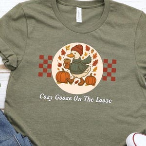 May include: Olive green t-shirt featuring a cartoon goose wearing a hat and sweater, holding a coffee cup, and walking near pumpkins. The text "Cozy Goose On The Loose" is below the graphic. The shirt is shown with blue jeans and white sneakers.