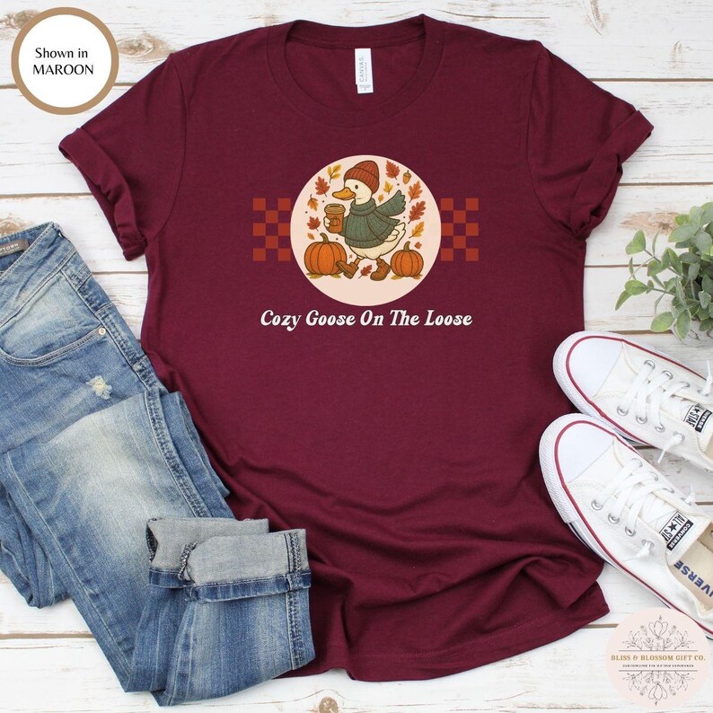 Puede incluir: Camiseta granate con un dibujo de un ganso de dibujos animados con sombrero y bufanda, sosteniendo una taza de caf&eacute;, rodeado de calabazas y hojas de oto&ntilde;o. El texto "Cozy Goose On The Loose" est&aacute; debajo. La camiseta se combina con vaqueros azules y zapatillas blancas.