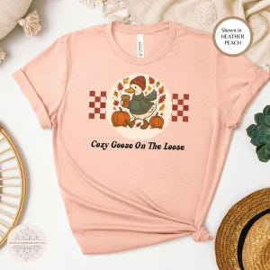 Puede incluir: Camiseta color melocot&oacute;n con un gr&aacute;fico de un ganso con sombrero y su&eacute;ter, sosteniendo una taza de caf&eacute; y calabazas. El texto "Cozy Goose On The Loose" est&aacute; debajo. La camiseta se muestra en Heather Peach.