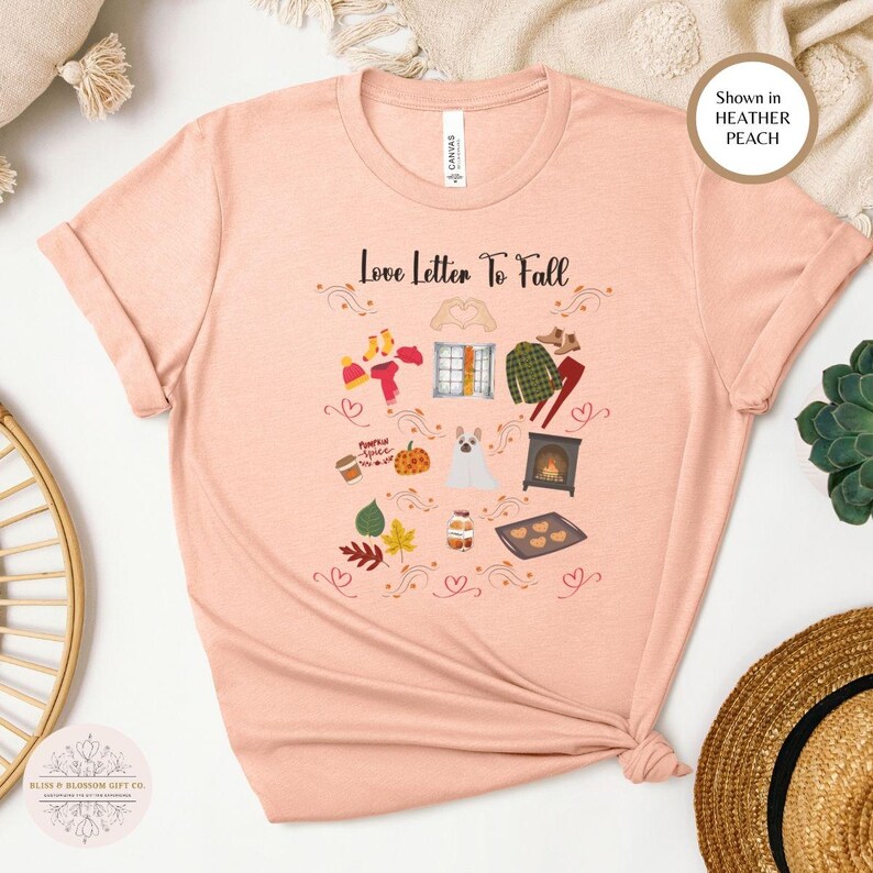 May include: A peach-colored t-shirt featuring a fall-themed graphic. The design includes the text 'Love Letter To Fall' along with illustrations of autumn elements like leaves, a pumpkin, a dog, a fireplace, and baked goods. The shirt is shown in a relaxed fit.