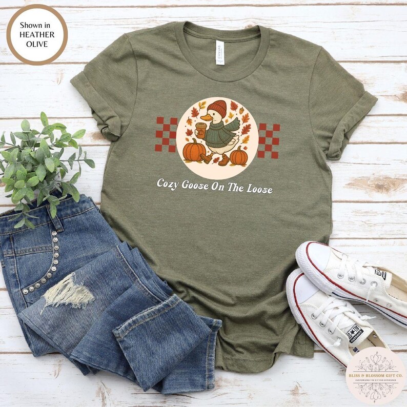 May include: Olive green t-shirt featuring a cartoon goose wearing a hat and sweater, holding a coffee cup, and walking near pumpkins. The text "Cozy Goose On The Loose" is below the graphic. The shirt is shown with blue jeans and white sneakers.