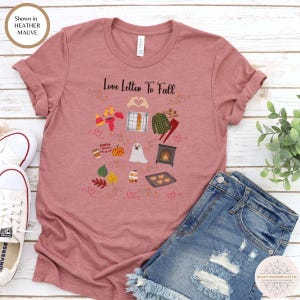 以下が含まれることがあります： ヘザーモーヴ色のTシャツで、中央に「Love Letter To Fall」という文字と、ゴースト、カボチャ、コーヒーカップ、チェック柄のシャツ、暖炉、ハートの形など、秋をテーマにした様々なイラストがグラフィックデザインとしてプリントされています。