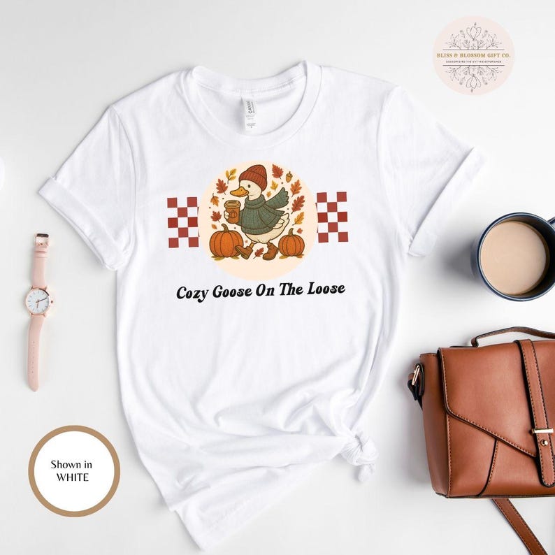 May include: White t-shirt featuring a cartoon goose wearing a hat and scarf, holding a coffee cup, and standing near pumpkins. The text "Cozy Goose On The Loose" is printed below the graphic. The shirt is shown in white.