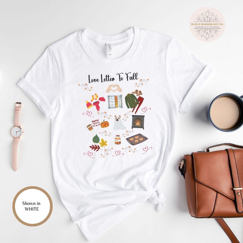 May include: White t-shirt with the text "Love Letter To Fall" and fall-themed illustrations. The design includes images of socks, a window, a plaid shirt, a pumpkin, a dog, a fireplace, and baked goods. A watch, a purse, and a coffee cup are also visible.