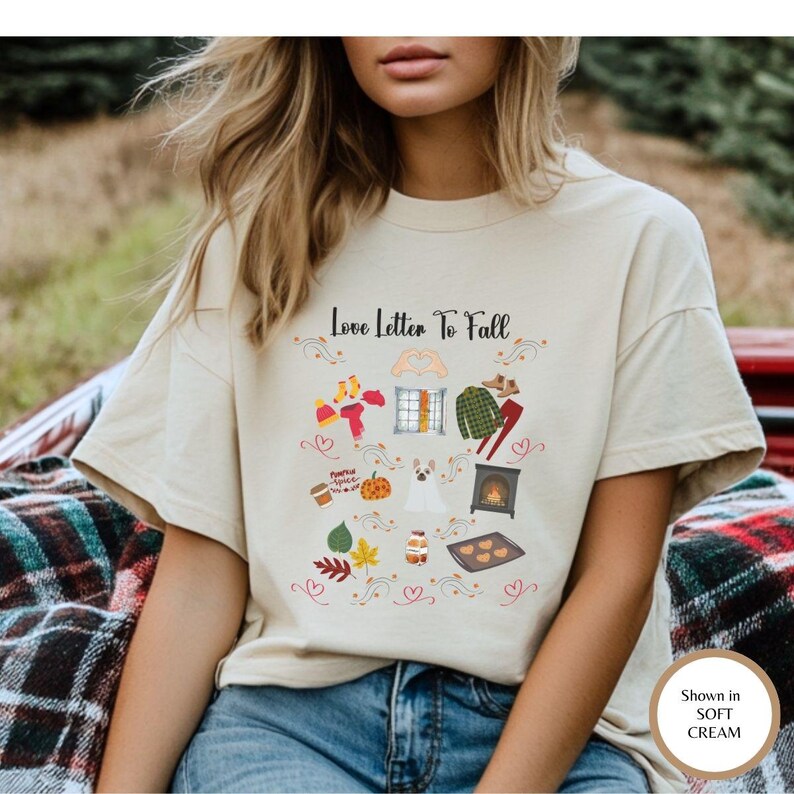 以下が含まれることがあります： Love Letter To Fallというテキストと、窓、チェック柄のシャツ、カボチャ、犬、暖炉、秋の葉、コーヒーカップ、季節の衣類などのカラフルなイラストが描かれた、ソフトクリーム色のTシャツ。