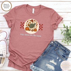 Puede incluir: Camiseta color malva jaspeado con un dise&ntilde;o de un ganso con sombrero y bufanda, sosteniendo una taza de caf&eacute; y calabazas. El texto "Cozy Goose On The Loose" est&aacute; debajo.
