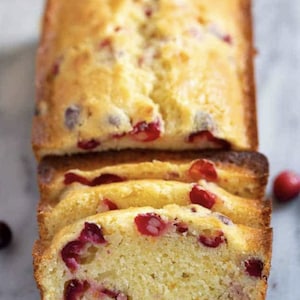 May include: A loaf of cranberry bread sliced open, revealing a light yellow crumb with red cranberries throughout. The bread is baked in a loaf pan and is ready to be served.