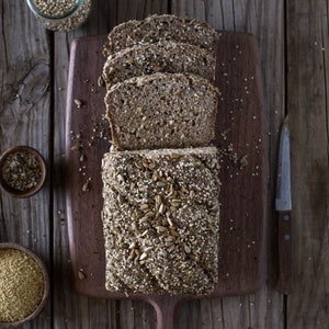 May include: A loaf of multigrain bread sliced on a wooden cutting board. The bread is covered in seeds, including sunflower seeds, sesame seeds, and flax seeds.