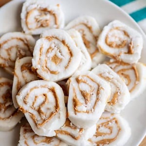May include: A white plate piled high with peanut butter and coconut roll-ups. The rolls are cut into bite-sized pieces and have a swirl of peanut butter in the center.