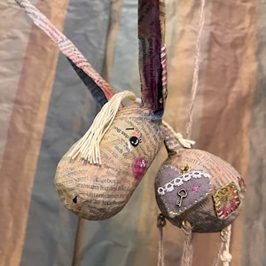 May include: Handmade paper mache unicorn and ball ornaments. The unicorn has a cream-colored mane and a pink cheek. The ball ornament has a heart-shaped patch with a key charm. Both are made from newspaper.