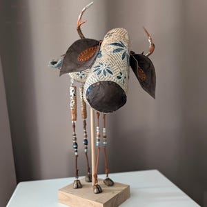 Könnte beinhalten: Eine dekorative Skulptur eines hirschartigen Wesens mit einem Kopf aus bedrucktem Text und blauen Blumenmustern, dunkelbraunen Lederohren und kleinen Geweihen. Der Körper wird von vier dünnen Beinen mit Perlenakzenten und Hufen getragen.