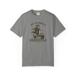 May include: A grey short-sleeve t-shirt featuring a graphic of a raccoon wearing goggles and a scarf while riding a green scooter. The text above the raccoon reads OFF TO CAUSE A and the text below the raccoon reads KERFUFFLE in dark lettering.